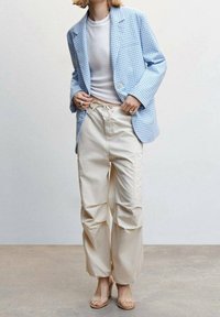 Blazer in cotone a scacchi blu chiaro sopra una maglietta bianca a coste, abbinato a pantaloni cargo beige. L'outfit presenta una vestibilità rilassata e dettagli con vita regolabile.