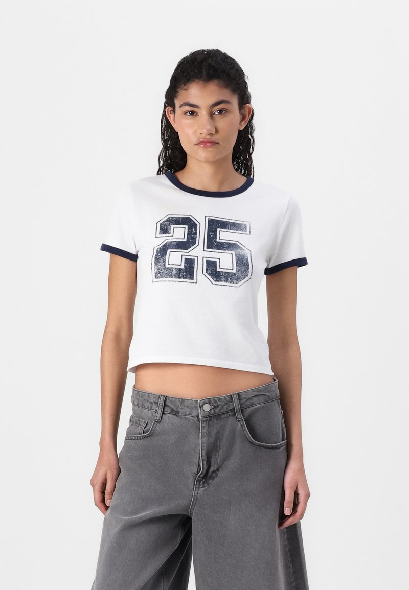 White cropped t-shirt with navy blue trim, featuring a distressed “25” graphic on the front. Paired with gray wide-leg pants.