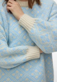 Personne portant un pull en tricot bleu clair et crème à motifs avec des poignets côtelés, les bras croisés et une main touchant le cou.