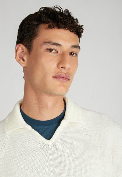 Helle cremefarbene Strickjacke mit V-Ausschnitt und flachem Kragen, über einem dunkelblauen Hemd getragen, das dezent strukturierte Details aufweist.