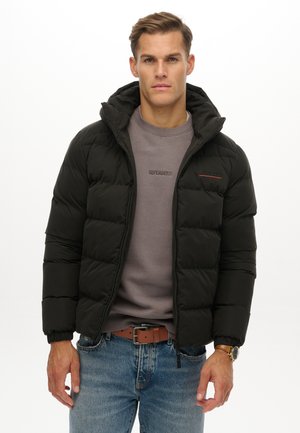 Młody mężczyzna ubrany w czarną pianę z kapturem, szarą bluzę, niebieskie dżinsy, brązowy pasek i zegarek na nadgarstku, stojący na białym tle.