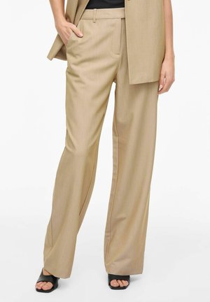 Bézs, széles szárú nadrág oldalzsebekkel, övbújtatóval és elülső hajtással, fekete nyitott orrú szandállal és hozzáillő bézs blézerrel viselve.