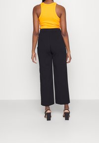 Pantalon noir à jambes larges taille haute, associé à un débardeur orange ajusté. La tenue présente une texture lisse et une coupe structurée.
