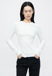Fehér hosszú ujjú felső bordázott textúrával, fekete gombdíszekkel az oldalán, kerek nyakkivágással és testhez simuló szabással. Fekete nadrággal párosítva.
