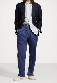 Blazer de marina de doble pecho sobre una camisa de botones a rayas, complementado con pantalones azules con cinturón y zapatillas de color claro.