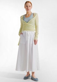 Kvinde med rødt hår iført bleggul wrap-striktrøje over grå tanktop, lang hvid nederdel med lommer og lyseblå balletsko, stående.