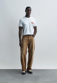 Άνδρας στέκεται με το ένα χέρι στην τσέπη, φοράει λευκό μπλουζάκι με κόκκινο λογότυπο, καφέ παντελόνι με φαρδιά ποδία και μαύρα-μπεζ παπούτσια.