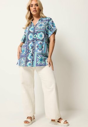 Mujer con una camisa azul de manga corta con patrón suelto, pantalones blancos de pierna ancha y sandalias de plataforma beige, de pie frente a un fondo liso.