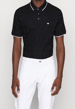 Man draagt een zwarte polo met korte mouwen, voorzien van een subtiel tekstpatroon en witte bies, gecombineerd met witte broek met knoopsluiting aan de voorkant.