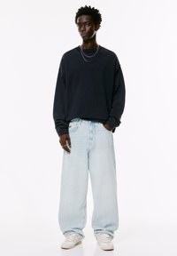 Ung mand iført en sort oversized sweatshirt, lyseblå vide jeans, hvide sneakers og to sølv kæder, der står med hænderne i lommen.