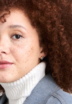 Kolczyki w kształcie serca z wielokolorowymi cyrkoniami. Wykonane z metalu, eksponowane obok swetra typu golf w neutralnym kolorze i dżinsowej kurtki.