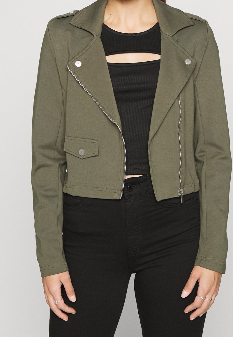 Giacca cropped verde oliva con hardware argento, dotata di una zip asimmetrica, colletto a punte e una tasca frontale. Abbinata a un top nero con ritagli.
