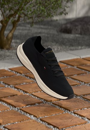 Black knit sneakers with a textured surface, cream midsole, and black outsole. Features dark laces and a logo on the tongue.