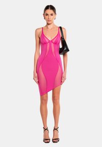 Rotes Netzkleid mit passender Silhouette, das spaghettiähnliche Träger, einen tiefen V-Ausschnitt und einen asymmetrischen Saum aufweist. Dazu eine schwarze Schultertasche und Riemchensandalen.