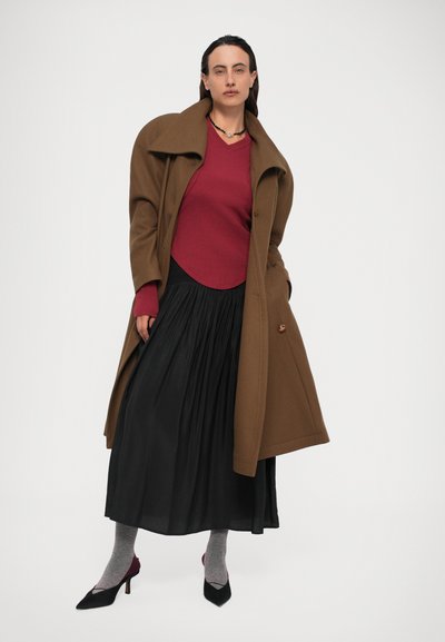 Donna che indossa un cappotto oversize marrone, una maglietta rossa a maniche lunghe, una gonna nera plissettata, calze grigie e tacchi neri su uno sfondo semplice.