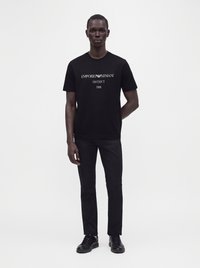 Άνδρας που φοράει μαύρο μπλουζάκι Emporio Armani με κείμενο, μαύρο παντελόνι και μαύρα αθλητικά παπούτσια, στέκεται πάνω σε απλό λευκό φόντο.