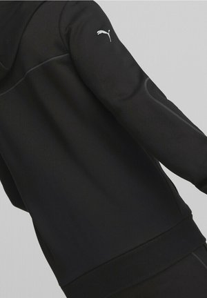 Achteraanzicht van een persoon die een zwarte Puma-jas draagt met een wit Puma-logo op de bovenarm, tegen een neutrale achtergrond.