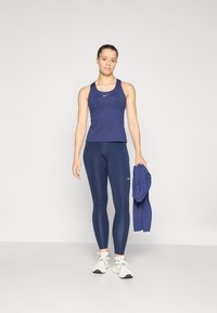 Tamnoplavi sportski top bez rukava i tajice, prilagođenog dizajna, glatke teksture. Nike logo na oba komada. Model drži usklađenu jaknu, nosi bijele tenisice.