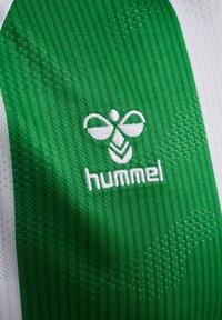 Zielona koszulka sportowa z teksturowanej tkaniny, białym haftowanym logo "hummel" oraz charakterystycznym wzorem w różnych odcieniach zieleni.