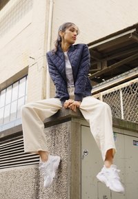 Veste bomber matelassée marine, t-shirt blanc, pantalons larges beiges et baskets blanches. Design minimaliste avec des matériaux texturés et des couleurs contrastées.