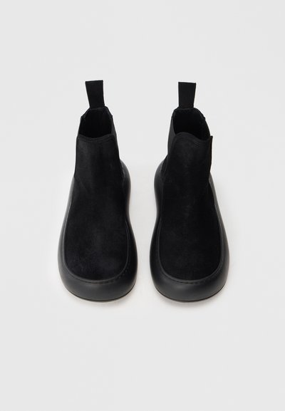 Schwarze Knöchelstiefel aus Wildleder mit Gummisohle. Verfügen über eine glatte Oberfläche und eine Zuglasche für ein einfaches An- und Ausziehen, ohne sichtbare Verschlüsse.