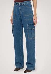 Modré denimové cargo kalhoty s uvolněným střihem, velkými bočními kapsami a vybledlou texturou. Obsahují poutka na opasek a přední zapínání na knoflík.