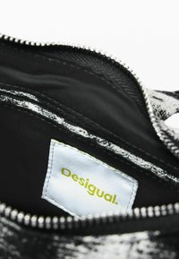 Wnętrze torby w czarno-białe wzory z silverowym zamkiem i zieloną metką "Desigual" na białej etykiecie. Gładka powierzchnia, materiał syntetyczny.