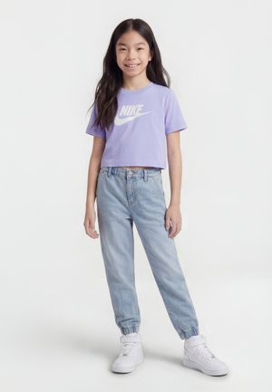 Dievča stojace a usmievajúce sa, oblečené v fialovom krátkom tričku Nike, svetlomodrých džínsoch jogger a bielych vysokých teniskách na bielom pozadí.