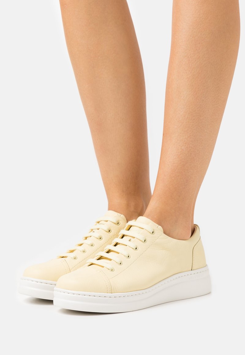 Jambes portant des baskets basses beiges avec des semelles blanches épaisses, debout sur une surface blanche sur un fond uni.