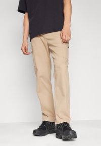 Man in beige cargobroek, zwarte sneakers en een zwart t-shirt, staande tegen een effen lichte achtergrond.