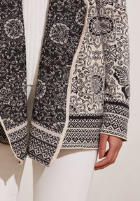 Svart och krämfärgad mönstrad kofta med intrikata mönster, texturerad tyg, öppen framsida och långa ärmar. Utsmyckad med geometriska kanter.