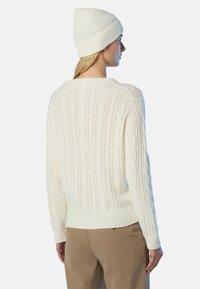 Donna che indossa un maglione bianco a trecce e un berretto coordinato, abbinati a pantaloni beige, in piedi di fronte a destra su uno sfondo semplice.