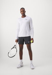 Camiseta deportiva blanca de manga larga con un botón en el cuello y un logo negro, combinada con pantalones cortos negros con bolsillos laterales, sosteniendo una pelota de tenis y una raqueta.