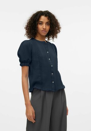 Kobieta z kręconymi włosami, ubrana w granatową bluzkę zapinaną na guziki z bufkami na rękawach i luźne szare spodnie na białym tle.