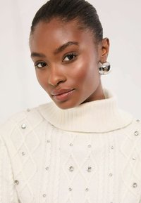 Fehér kötött pulóver magas gallérral, kábelmintás kötésű, elöl szórt kristálydíszítéssel. Ezüst karika fülbevalók.