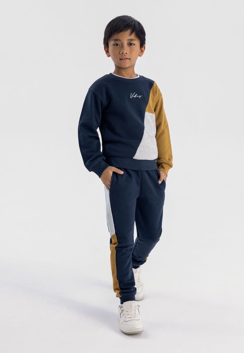 Mörkblå sweatshirt med grå och senapsgula lappar, med rund hals och logotyp. Matchande mörkblå joggers med sidopaneler i grått och senapsgult.