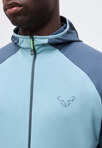 Κοντινό πλάνο ενός ατόμου που φοράει ένα φούτερ με φερμουάρ σε ανοιχτό και σκούρο μπλε χρώμα με μικρό ανάγλυφο λογότυπο ζώου στο στήθος.