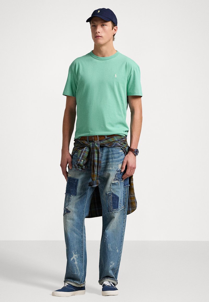 Mintgrünes T-Shirt mit Rundhalsausschnitt, zerschlissene Jeans mit Patches, karierte Bluse um die Taille gebunden, marineblaues Cap und blaue Sneaker.