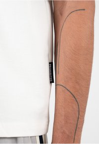 Hvitt bomull t-skjorte med en horisontal søm og et lite svart "FORBUDT" merke på siden; modellens arm med en enkel tatovering er synlig.