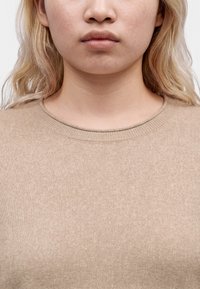 Jeune personne à la peau claire et aux cheveux blonds portant un pull en maille beige à col rond sur un fond uni.