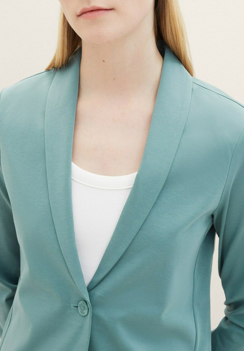 Lichte teal blazer met een geknipte revers, enkele knoopsluiting en een soepele stof. Draagt over een witte geribde tanktop.