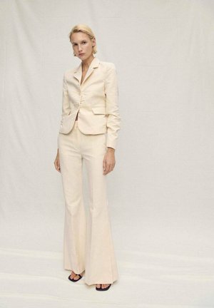 Blazer su misura beige chiaro con un bottone singolo, abbinato a pantaloni a campana. Tessuto liscio con una leggera lucentezza; il modello indossa sandali neri.