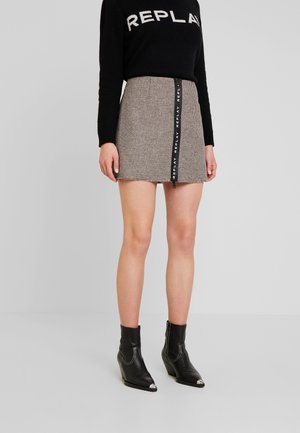 Femme portant un pull noir avec le texte "REPLAY", une mini-jupe à carreaux avec une fermeture éclair verticale "REPLAY" et des bottines noires.