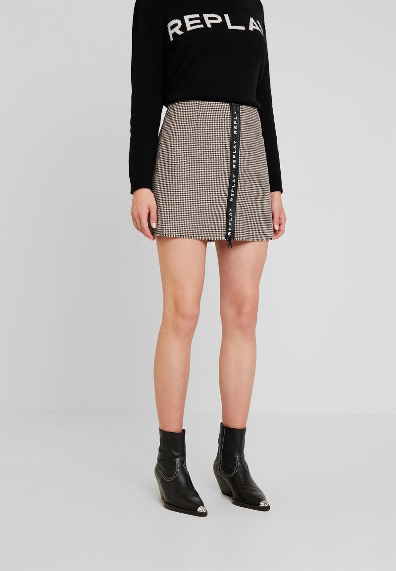 Femme portant un pull noir avec le texte "REPLAY", une mini-jupe à carreaux avec une fermeture éclair verticale "REPLAY" et des bottines noires.