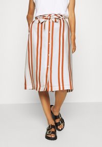 Jupe blanche à rayures verticales orange, longueur genou, avec une taille nouée, un devant boutonné et des poches latérales. Sandales noires à boucles.