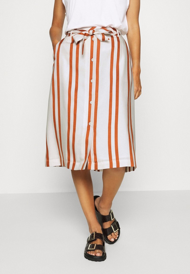 Jupe blanche à rayures verticales orange, longueur genou, avec une taille nouée, un devant boutonné et des poches latérales. Sandales noires à boucles.
