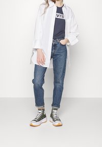 Camisa blanca de manga larga sobre una camiseta gráfica de color azul marino, combinada con vaqueros de mezclilla azules y zapatillas de caña alta verdes con cordones blancos y suelas de goma.