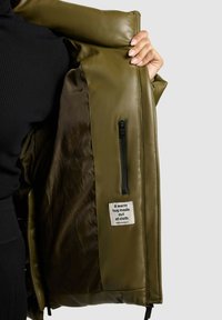 Olivgrüne Jacke mit glattem Lederaußenmaterial, ausgestattet mit einer Reißverschlusstasche und einem aufgenähten Etikett mit der Aufschrift "Eine warme Umarmung aus Stoff."