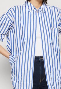 Camisa de botones a rayas azul y blanca con cuello y bolsillo en el pecho, confeccionada en un tejido ligero. Presenta mangas enrolladas y un corte relajado.
