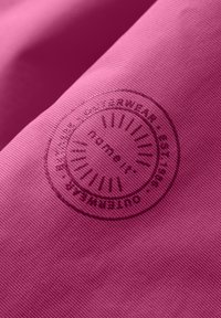 Pink textile featuring a textured surface, displaying a circular logo with embossed lettering and sunburst design.